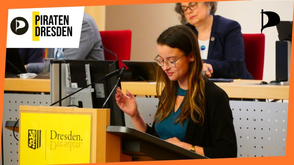 Stadträtin Anne Herpertz an einem Pult im Plenarsaal des Stadtrats Dresden. Um die Szene ein oranger Rahmen und zwei Logos der Piratenpartei.
