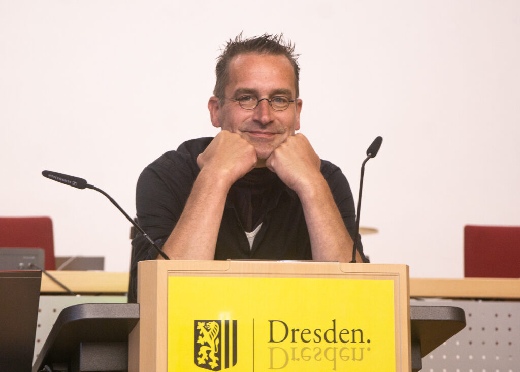 Dr. Martin Schulte-Wissermann, wie er mit den Ellebogen auf ein Rednerpult, auf dem das Logo der Stadt Dresden ist, in die Kamera lächelt.