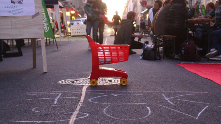 Ein Kindespielzeugswagen auf der Straße. Menschen sitzen auf der Straße. mit Kreide steht FCK SUV auf der Straße