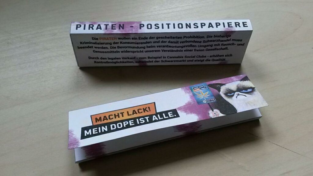 Zu sehen sind zwei Packungen Longpaper, "Positionspapiere" genannt. Auf der Hinterseite des einen ist Position der PIRATEN zu Pro-Legalisierung zu lesen. Auf der Vorderseite der anderen steht der Schriftzug "Macht Lack! Mein Dope ist alle.", daneben ein Bild von "Grumpy Cat", die ein Pro-Legalisierung Plakat des PIRATEN hoch hält.